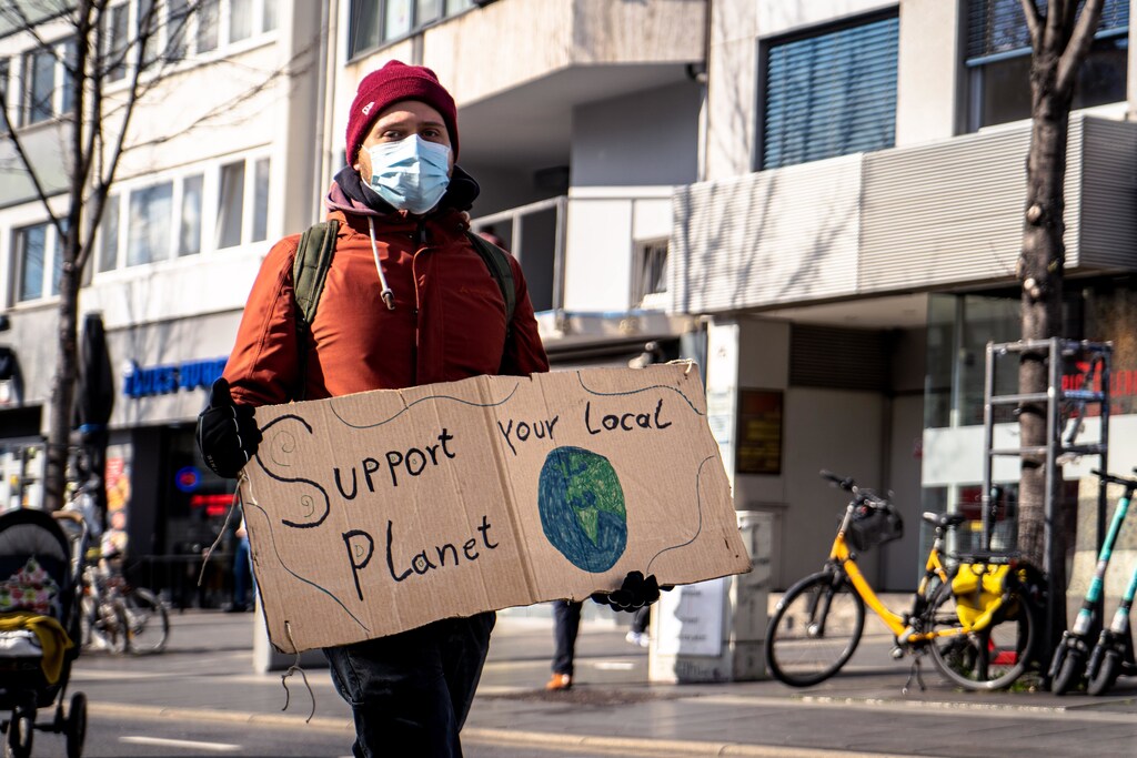 Man in red jacket and mask holding cardboard sign, "Support Your Local Planet": Employees can drive progress towards climate goals by insisting on a clear strategy on climate change from their employers.