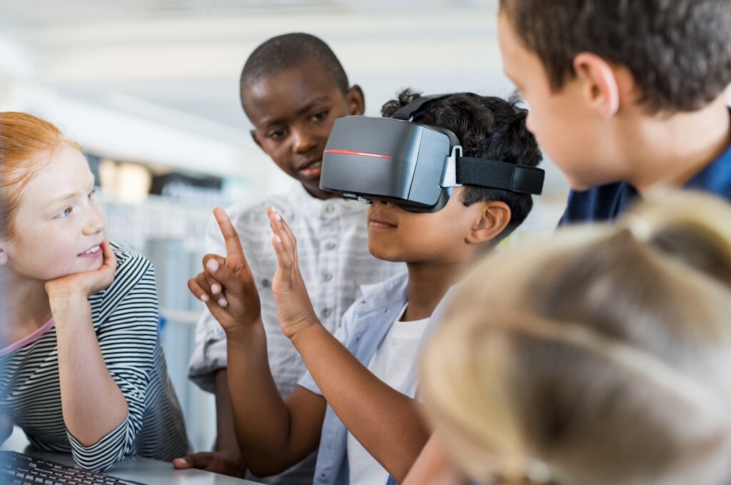 Mixed race child with vr virtual reality goggles in classroom with his friends. Multiethnic pupil having fun with virtual reality headset at elementary school. Happy boy gesturing while using VR headset in classroom.