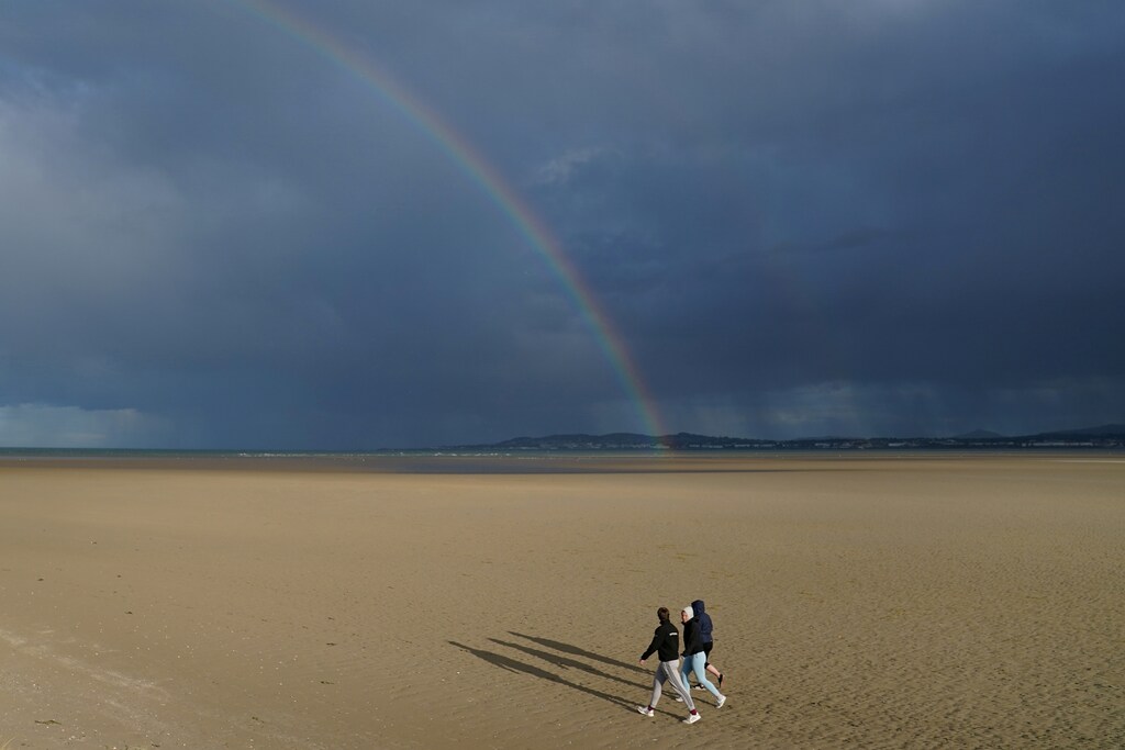 A rainbow and heavy rain are seen in the distance as people walk along an empty beach in sunshine, following the outbreak of the coronavirus disease (COVID-19), in Dublin, Ireland, June 5, 2020. REUTERS/Clodagh Kilcoyne - RC293H98WKRB
