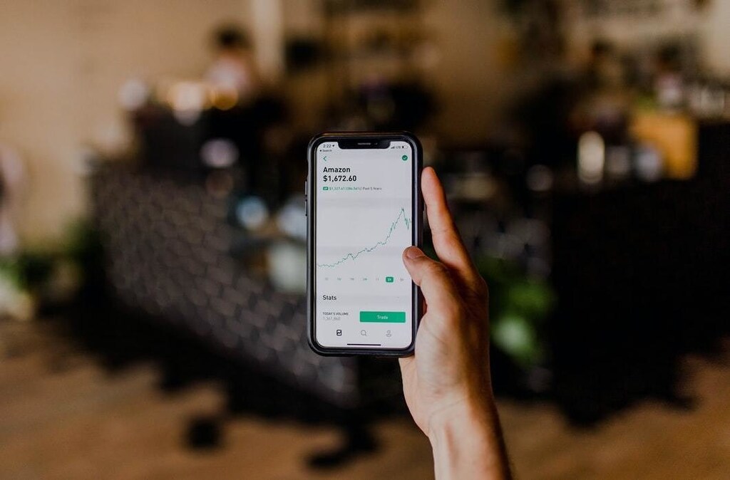 A man's hand holds a smart phone, showing the rising stock price of Amazon on its screen.