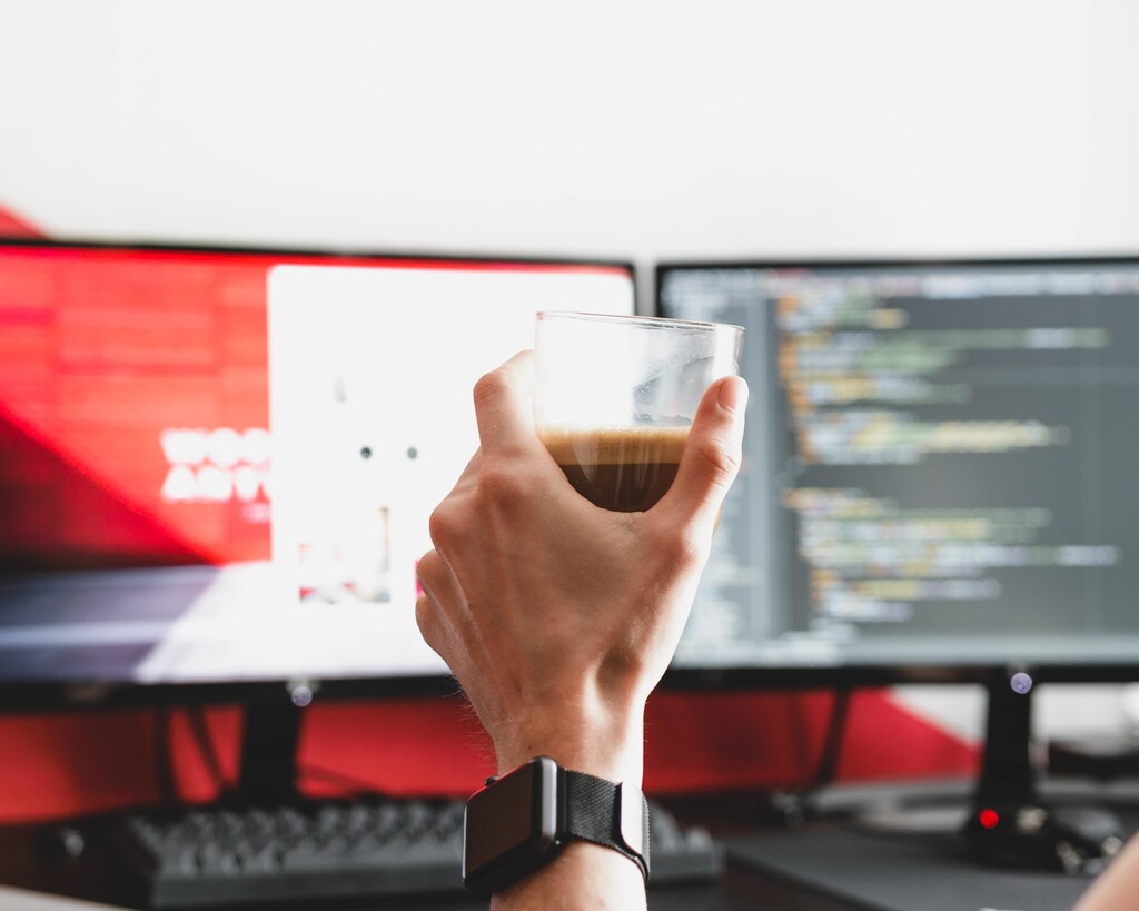 A hand holding a drink with two computer monitors in the background