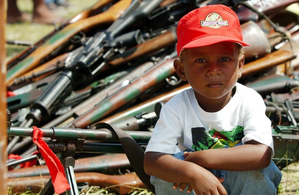 A young boy sits in front of weapons and ammunitions