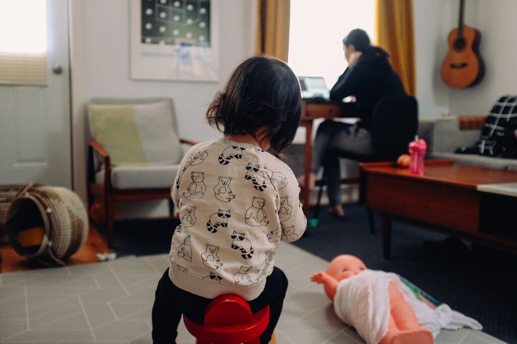 Una niña pequeña juega en primer plano mientras su madre está en una computadora en un blog sobre la economía del cuidado.