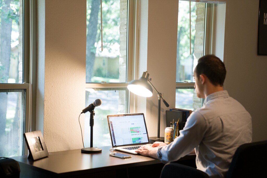 A man working from home, sat at a desk with a laptop and a microphone.