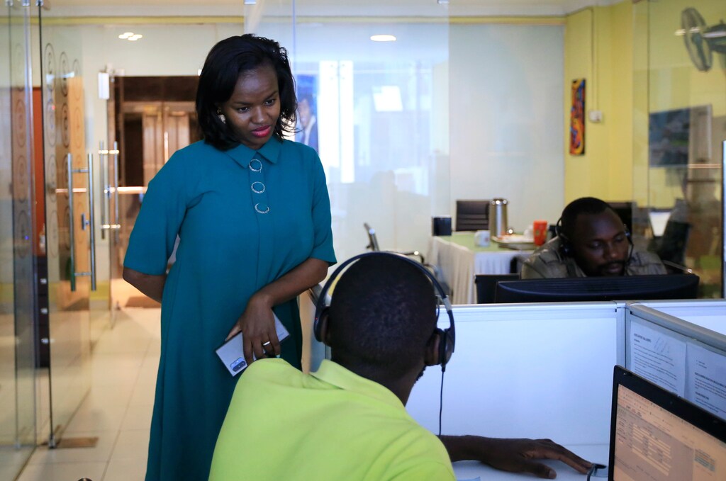 a woman talks to man sitting at a computer desk with a headset on