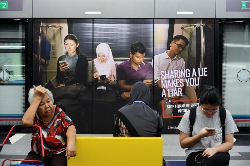 Commuters sit in front of an advertisement discouraging the dissemination of fake news, at a train station in Kuala Lumpur, Malaysia March 28, 2018. Picture taken March 28, 2018.