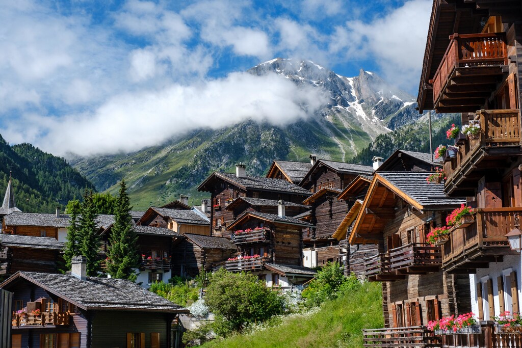 Brown wooden houses near green trees and a mountain under white clouds during daytime.