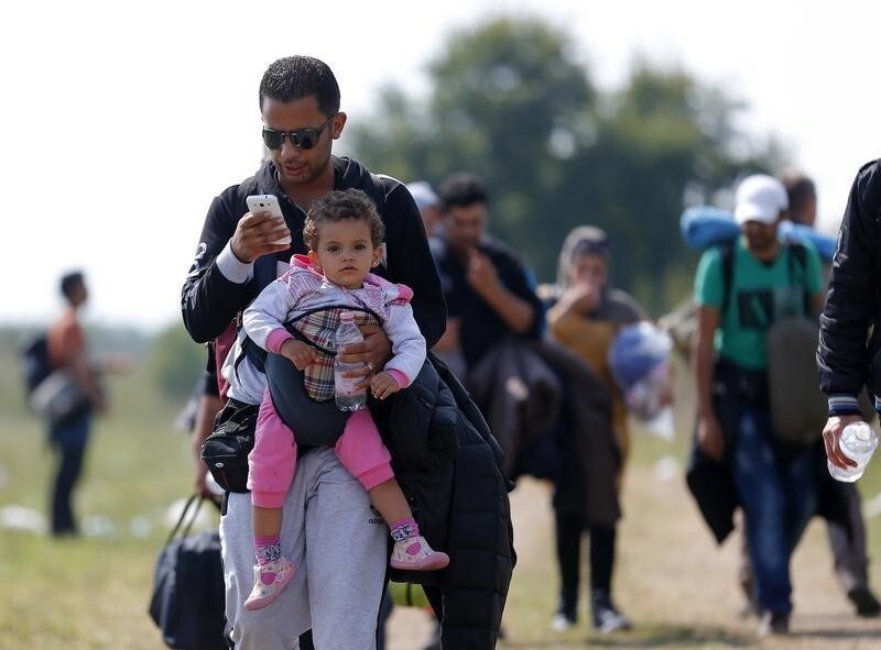 A migrant uses his mobile phone carrying his child as he walks through fields with others next to a collection point near the Serbian Hungarian border in Roszke, Hungary September 13, 2015. REUTERS/Laszlo Balogh