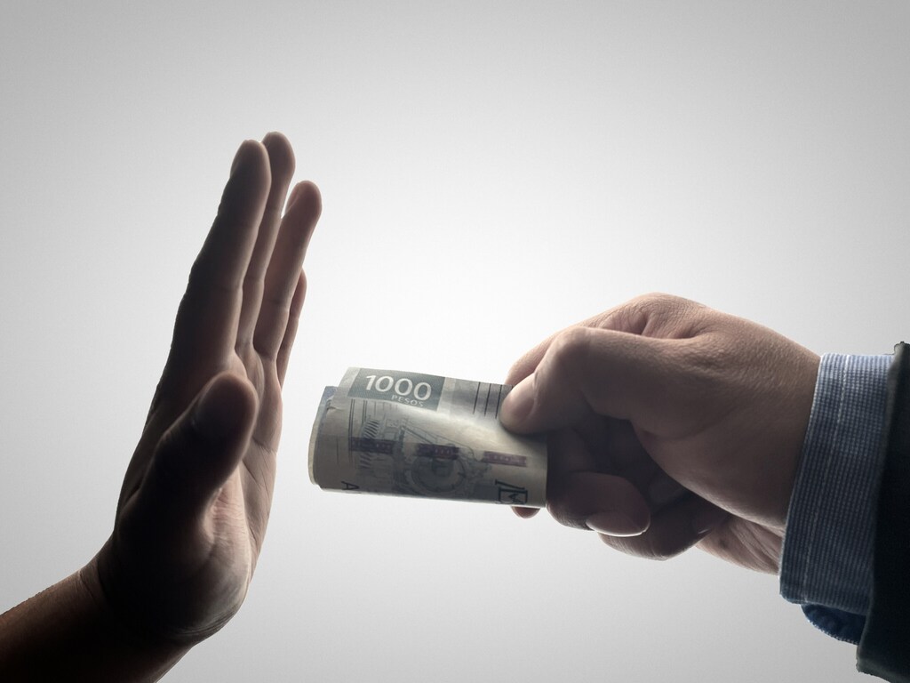 Una mano ofrece un fajo de billetes, mientras otra mano se levanta para decir rechazarlo.