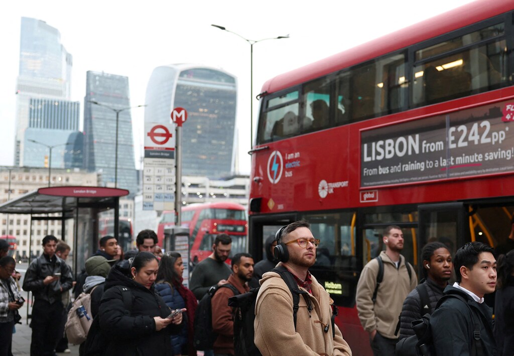 Fotografía de trabajadores esperando en una parada de autobús en London Bridge durante la hora pico de la mañana, con el paisaje urbano de Londres al fondo.