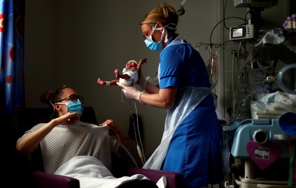 A nurse hands a new born baby to its mother while both wearing face masks.