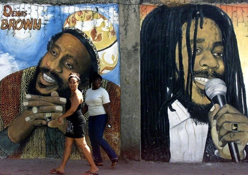 Jamaicans walk in front of a mural of reggae singers Denis Brown (L)and Bob Marley in Kingston February 17, 2002. Britain's Queen Elizabethand her husband, the Duke of Edinburgh, will visit Jamaica fromFebruary 18-20. REUTERS/Jorge SilvaKW - RP3DRHYPBFAA