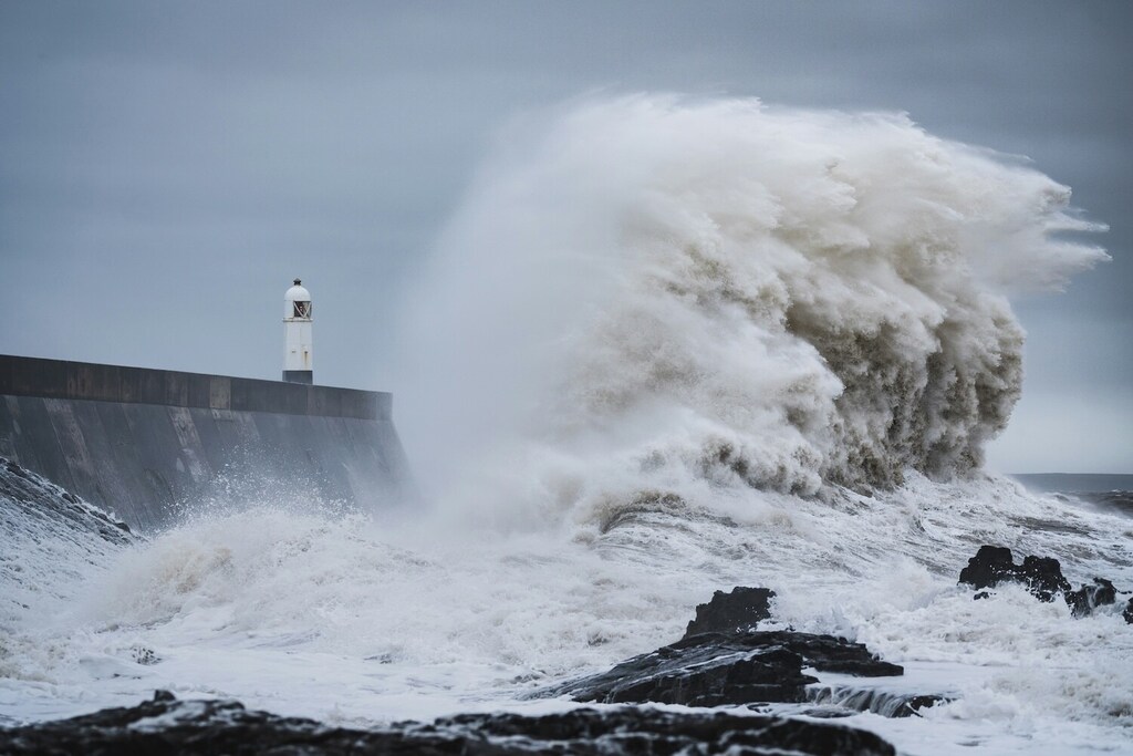 A massive wave crashing violently against a concrete sea wall, with a white lighthouse visible in the background under a gray sky.