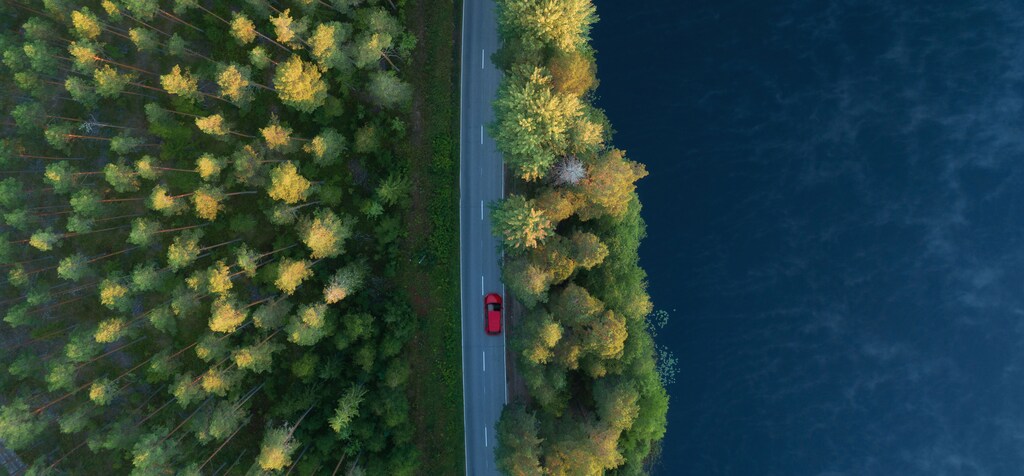 Aerial view of red car driving next to forests and body of water: Multiple powertrain technologies including hybrids will serve a cleaner tomorrow via electrification.