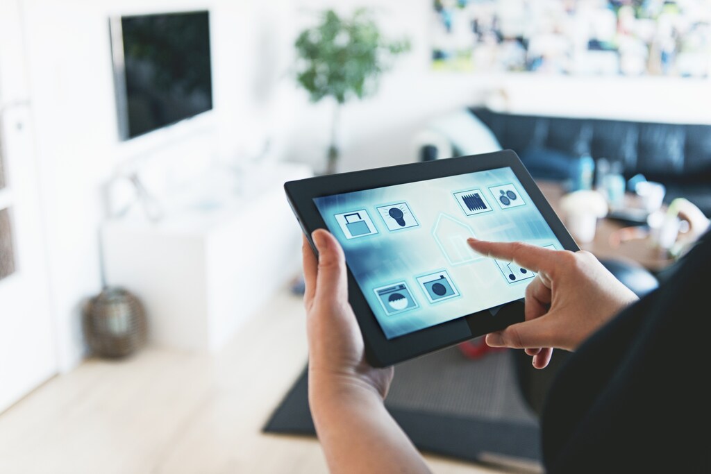 Connected devices: Tablet with an app controls several features in the home such as door lock, lights, washing machine, surveillance camera, music, water use and more. Woman stands in a living room and press the center of the tablet with an icon of a house.