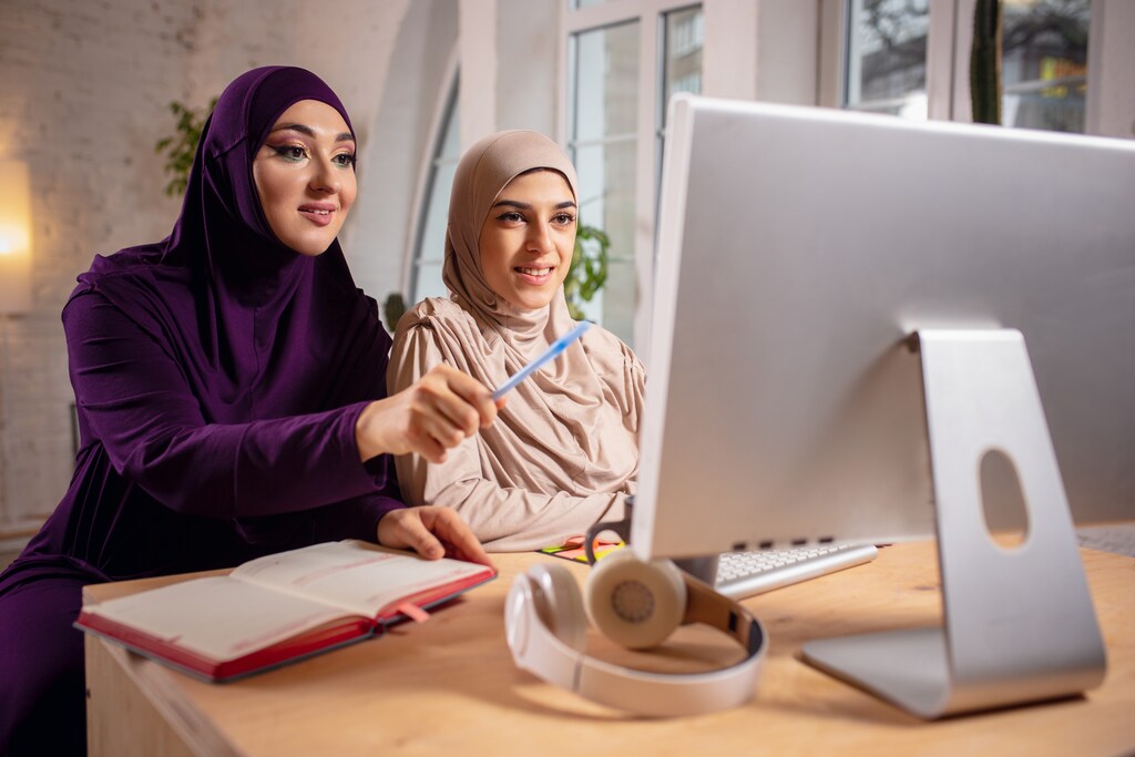 Pointing. Happy and young two muslim women at home during lesson, studying near computer, online education. Culture, traditions, modern people. Watching at PC monitor, shopping or talking.