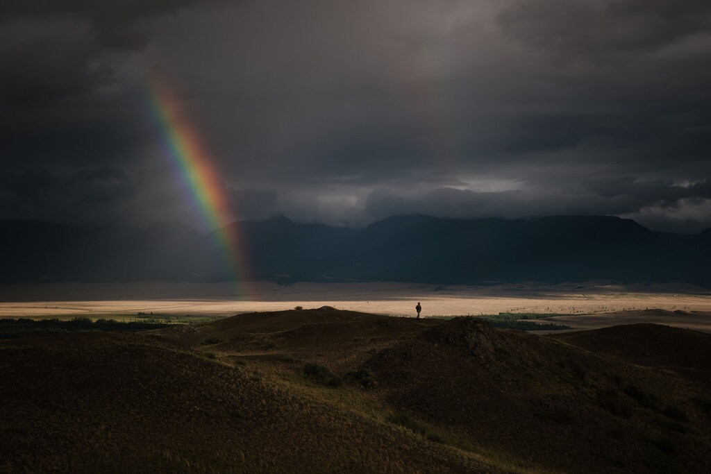 Breathtaking view of bright rainbow in dark sky with overcast above green mountain valley.