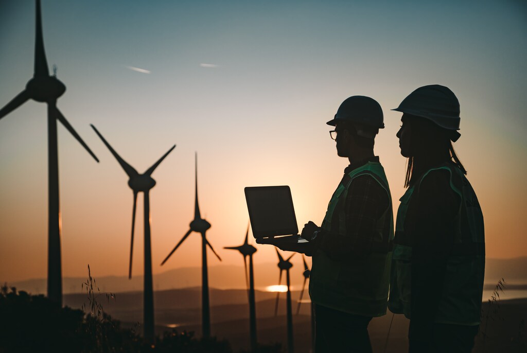 Climate transition plan. Silhouette shot of young electrical engineer woman and business man standing in front of wind turbines checking and working about technical problems and writes the results of measurements with laptop pc in wind power plant electric energy station. xxxl size taken with canon 5d mIV