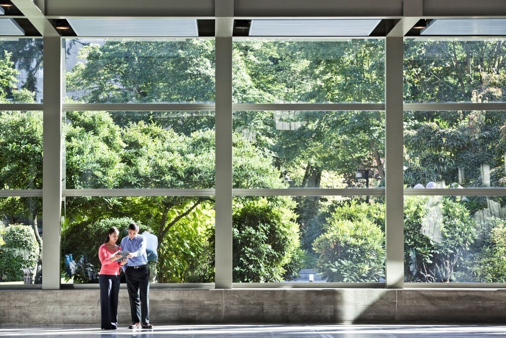 Two people in work clothes looking at a tablet in one of a growing number of sustainable buildings; green trees outside windows.