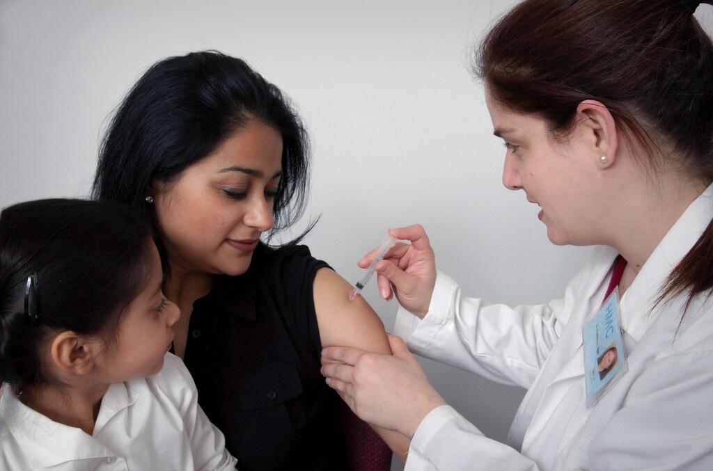 White female nurse injecting Asian woman with vaccine while child watches.