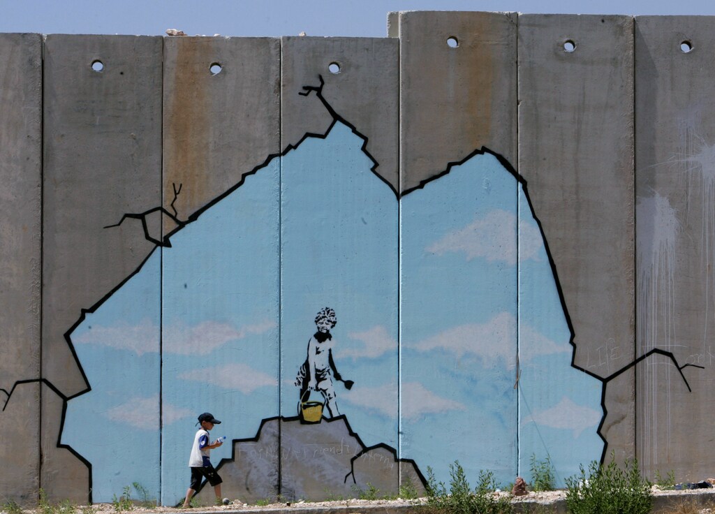 A Palestinian boy walks past a drawing by British graffiti artist Banksy near the Kalandia checkpoint in the West Bank. A Palestinian boy walks past a drawing by British graffiti artist Banksy, along part of the controversial Israeli barrier near the Kalandia checkpoint in the West Bank August 10, 2005. REUTERS/Ammar Awad - RTRK3ET
