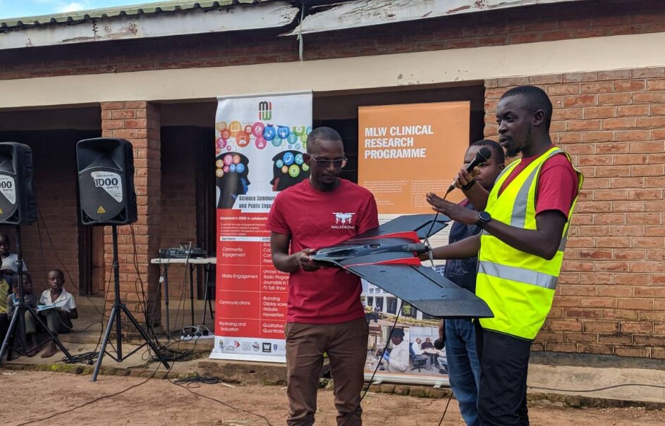 Patrick Kalonde, one of the drone pilots, speaks into a microphone as he briefs the community about the malaria project while his colleague Kennedy Zembere holds a drone in the Kasungu district in Malawi. 30 May 2020.