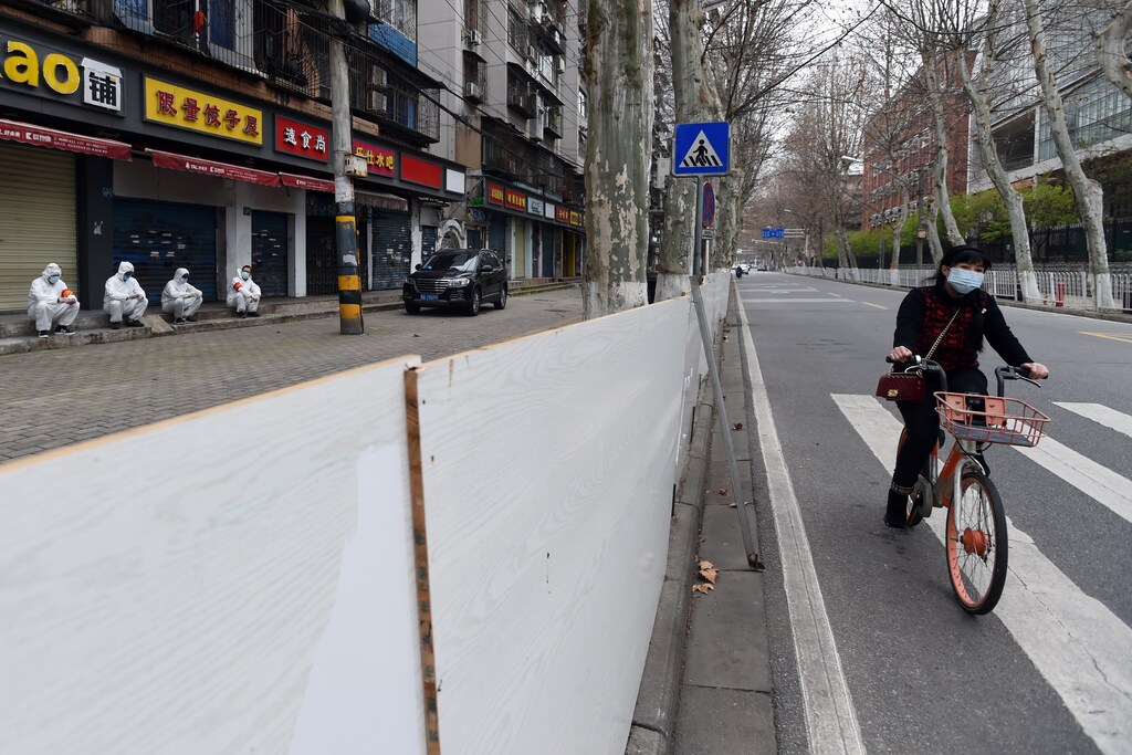 A woman wearing a face mask rides a shared bicycle past workers in protective suits who are resting in front of closed shops sealed off from the road, in Wuhan, the epicentre of the novel coronavirus outbreak, Hubei province, China March 6, 2020. REUTERS/Stringer CHINA OUT. - RC2CEF9NRA68