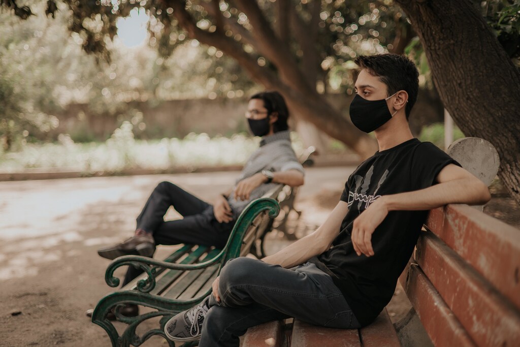 Two men sit on a bench, wearing masks.