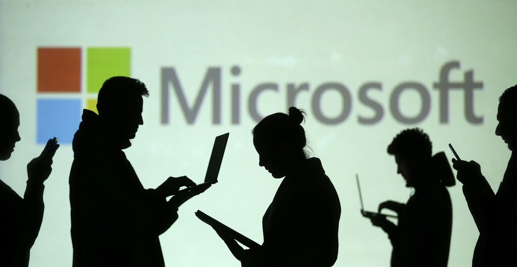 Silhouettes of laptop and mobile device users are seen next to a screen projection of Microsoft logo in this picture illustration taken March 28, 2018. REUTERS/Dado Ruvic/Illustration - RC1DBA871400