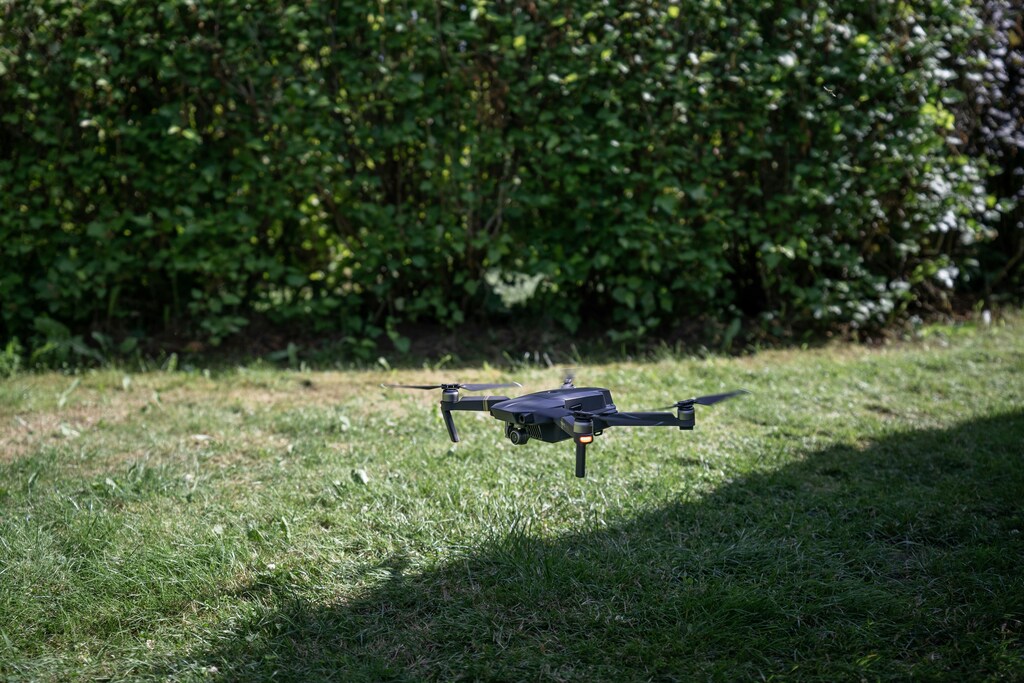 Grey quadcopter flying above the ground; green grass and hedge. Embodied AI