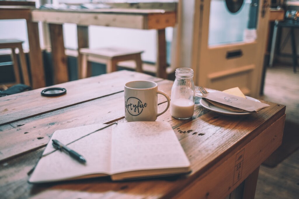 A writing journal and pen on a wooden table with mug, milk and cutlery