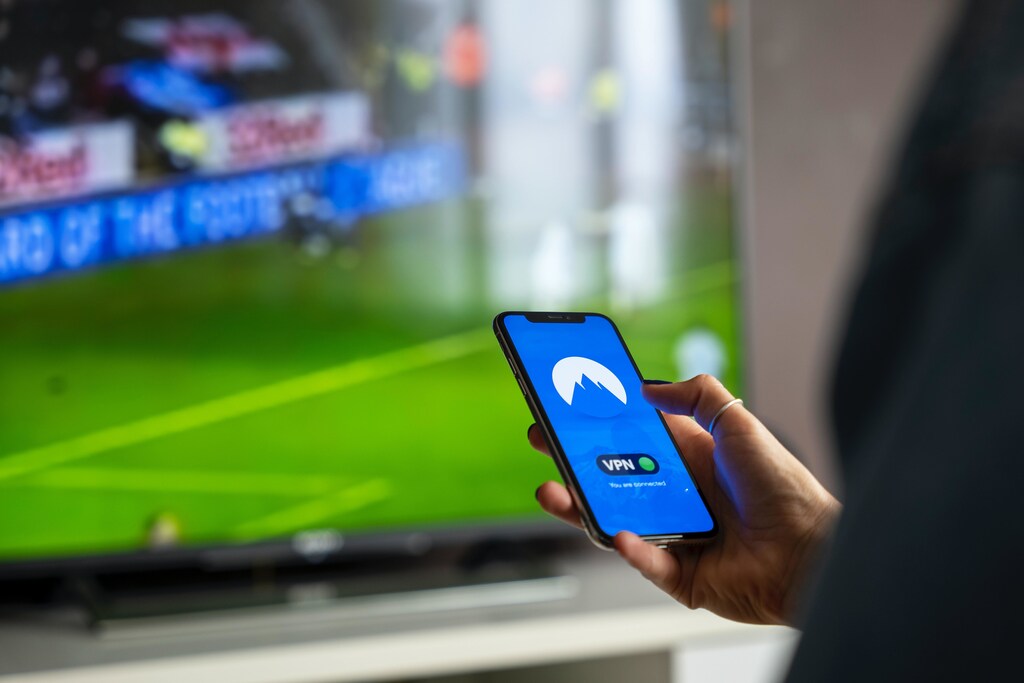 A person using a VPN on a mobile phone in front of a football game on a large TV screen.