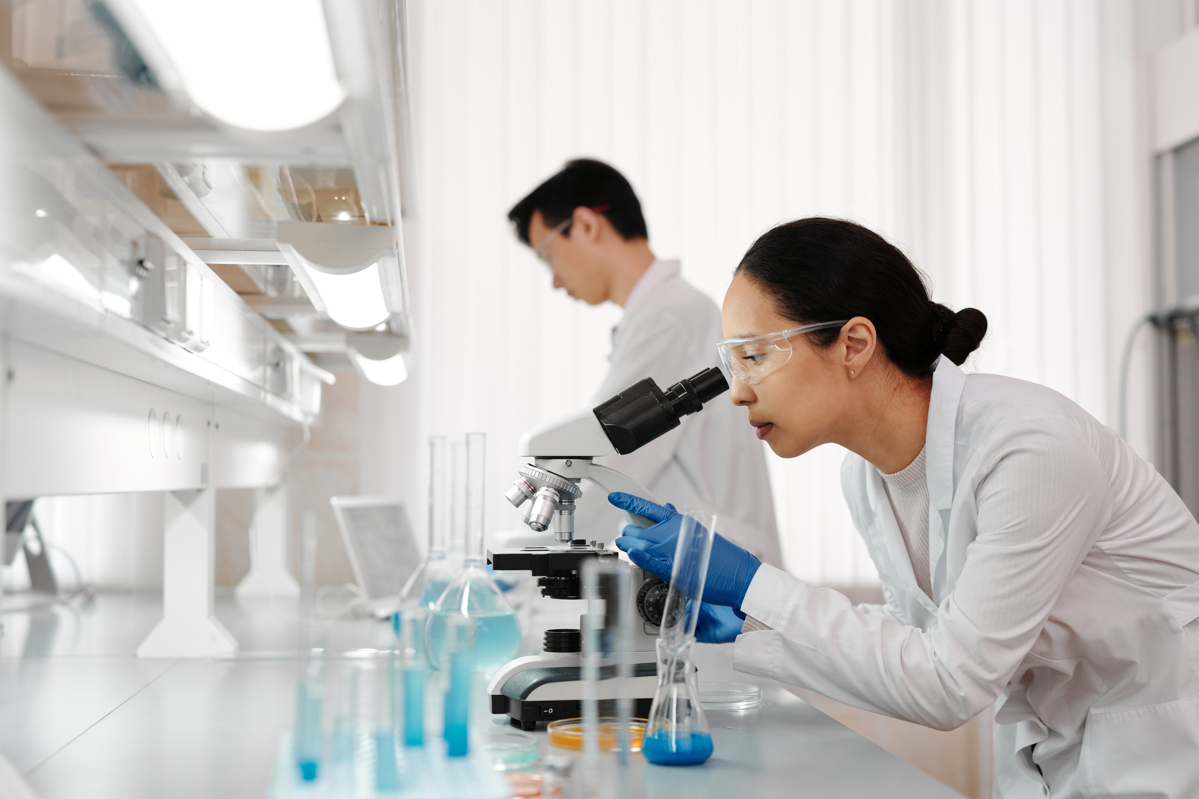 Woman in white lab coat looks through a microscope with colleague in background: Mistrust is preventing the use of AI in healthcare.