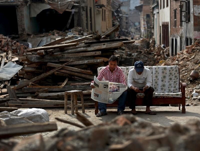 A man reads a newspaper as his friend looks at his mobile phone, as they sit next to collapsed houses on the outskirts of Kathmandu, Nepal, May 15, 2015.