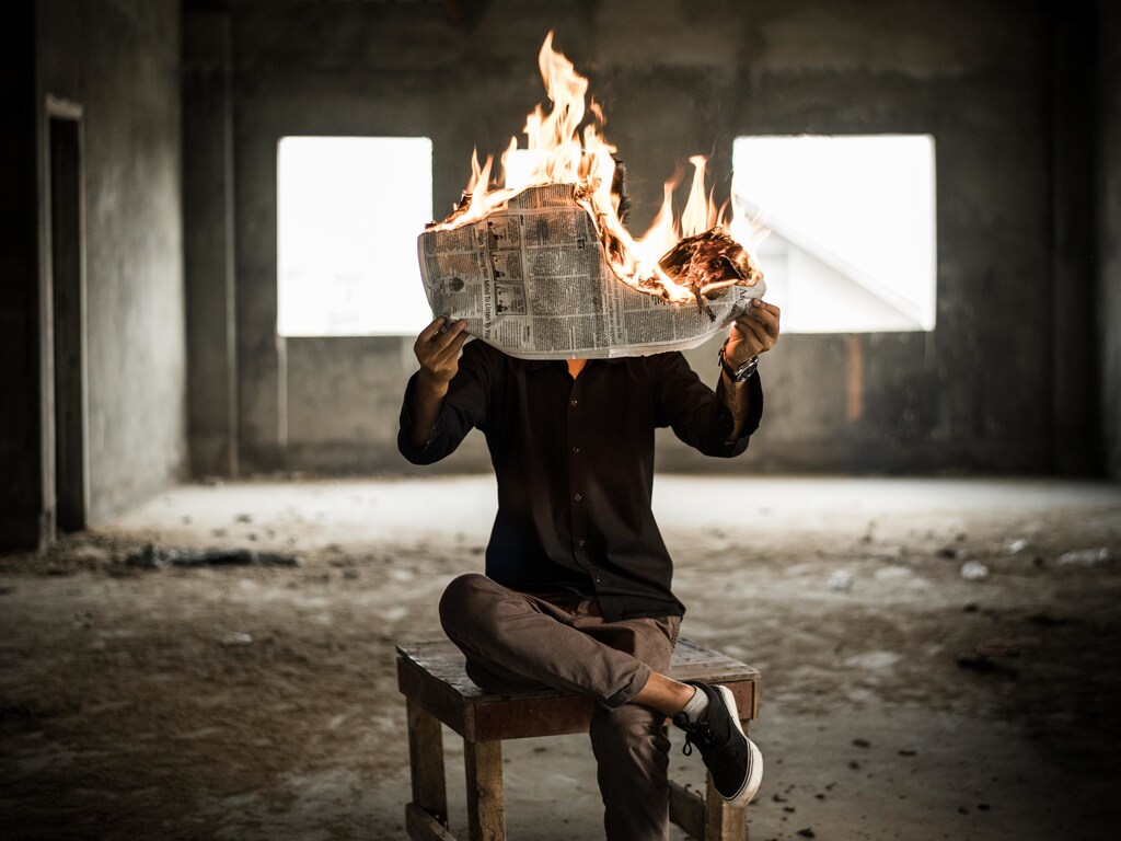 man sitting on chair holds a newspaper that's on fire