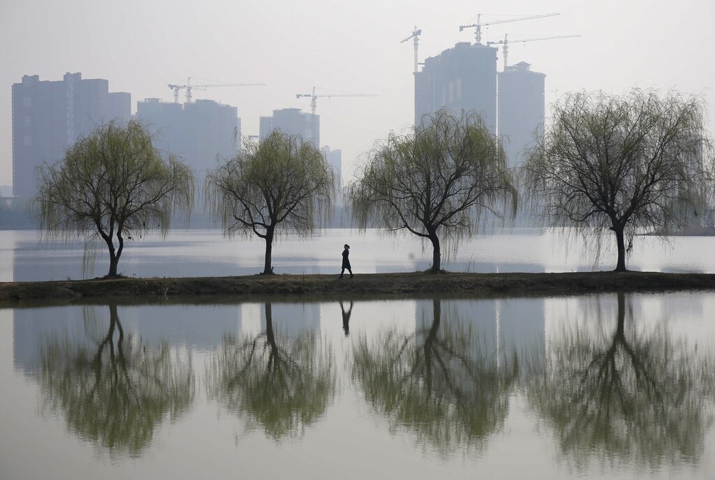 A woman walks past trees reflected on a lake in front of a construction site of a residential compound on a hazy day in Wuhan, Hubei province March 6, 2015.