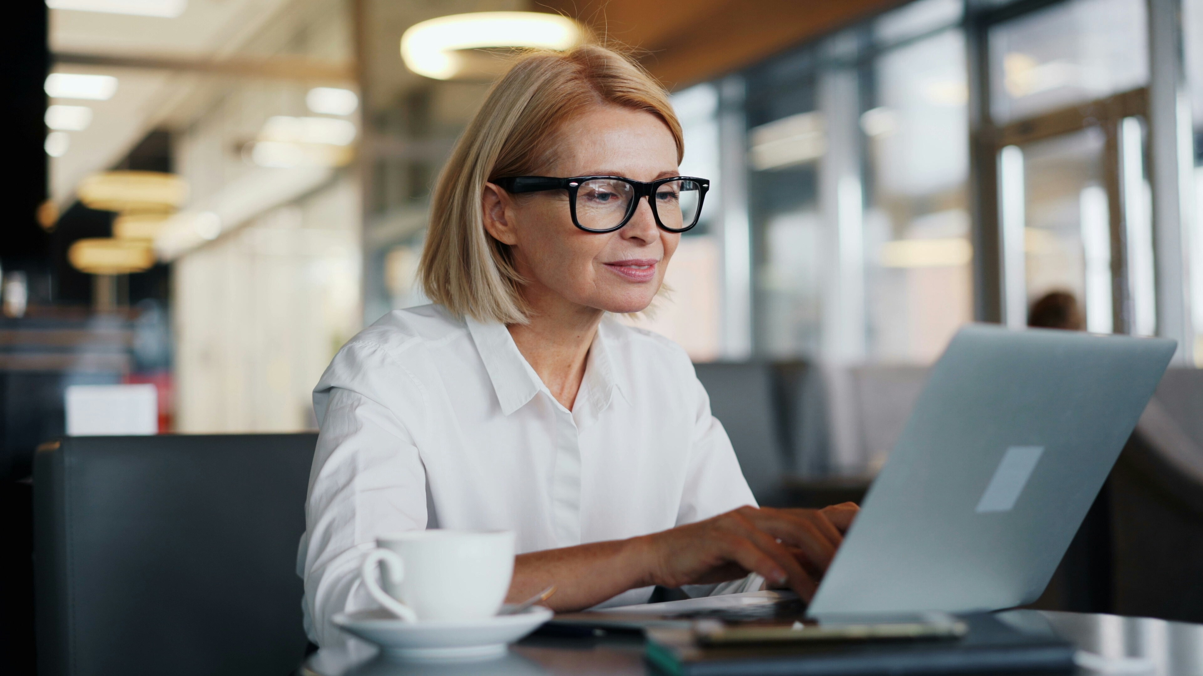A woman with blonde hair and glasses types at a computer, there is a cup on the table. Consumer spending across generations.