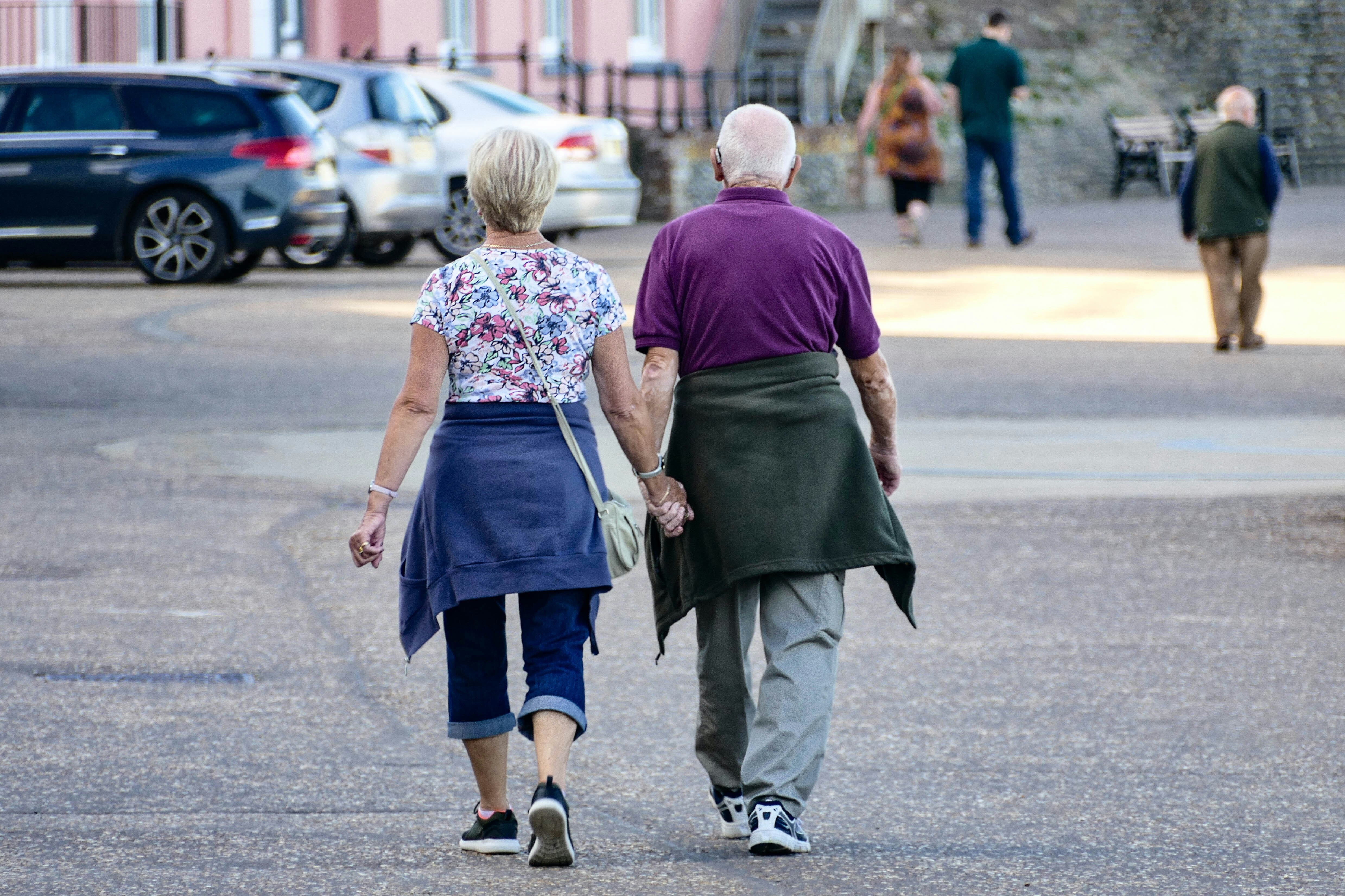 An older couple, one wearing a hearing aid, are seen holding hands and walking through a car park.