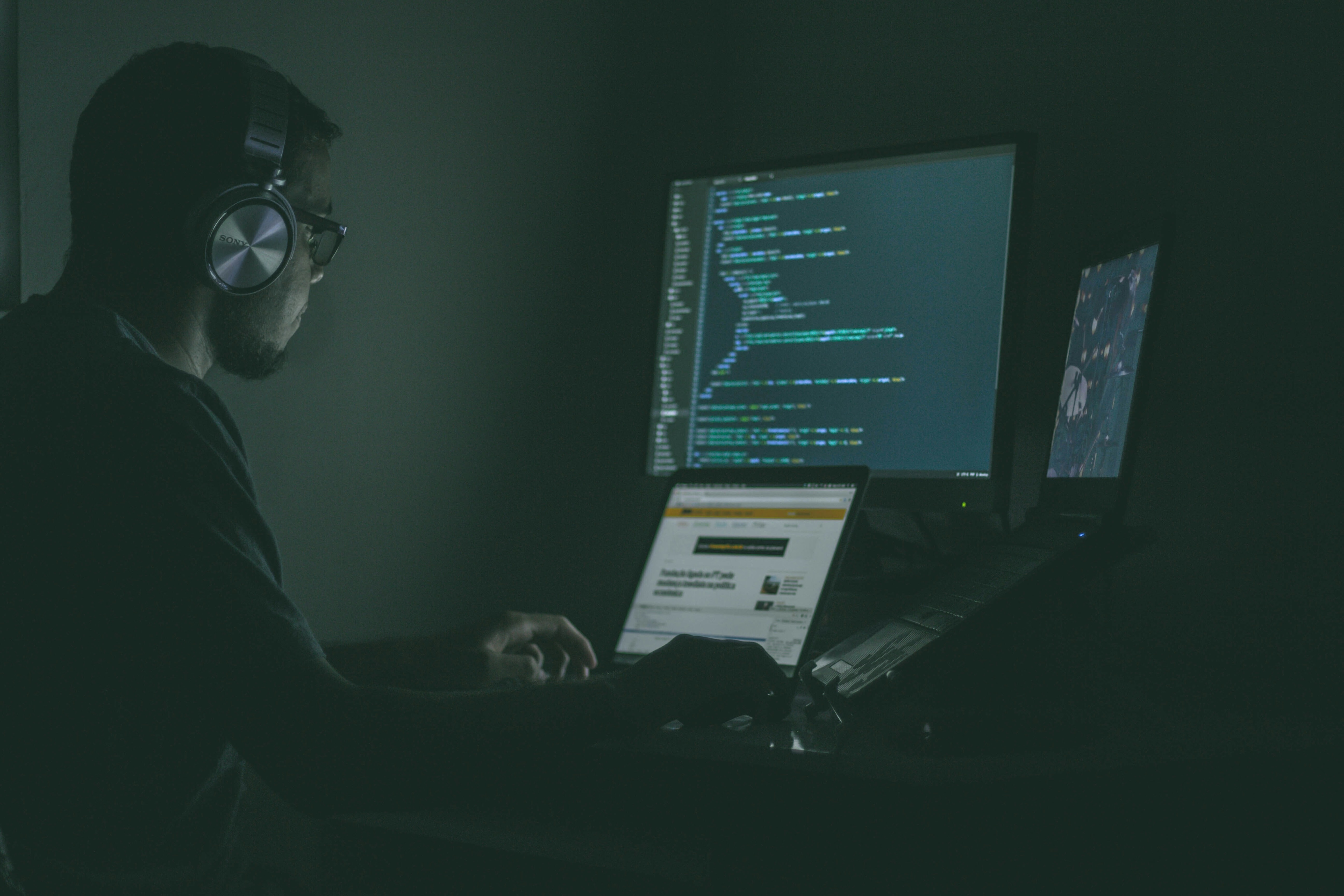 A man with headphones in front of several devices and montitors in a dark room: AI can help automate defences against cyber threats