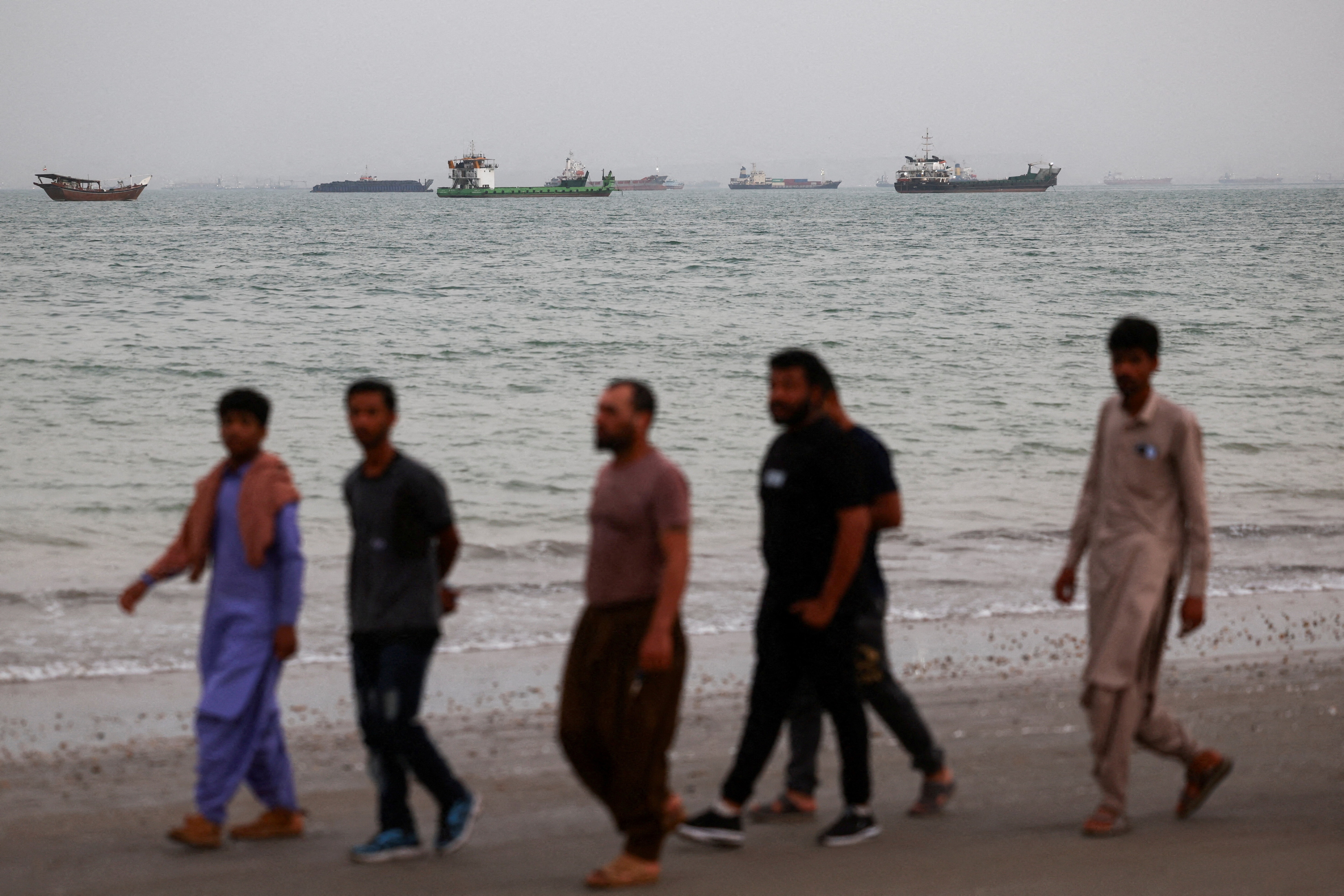 People walk on the beach, with vessels in the Strait of Hormuz visible near the beach of Bandar Abbas, Iran, May 22, 2026: The US-Iran war is creating a short-term brake on the energy transition