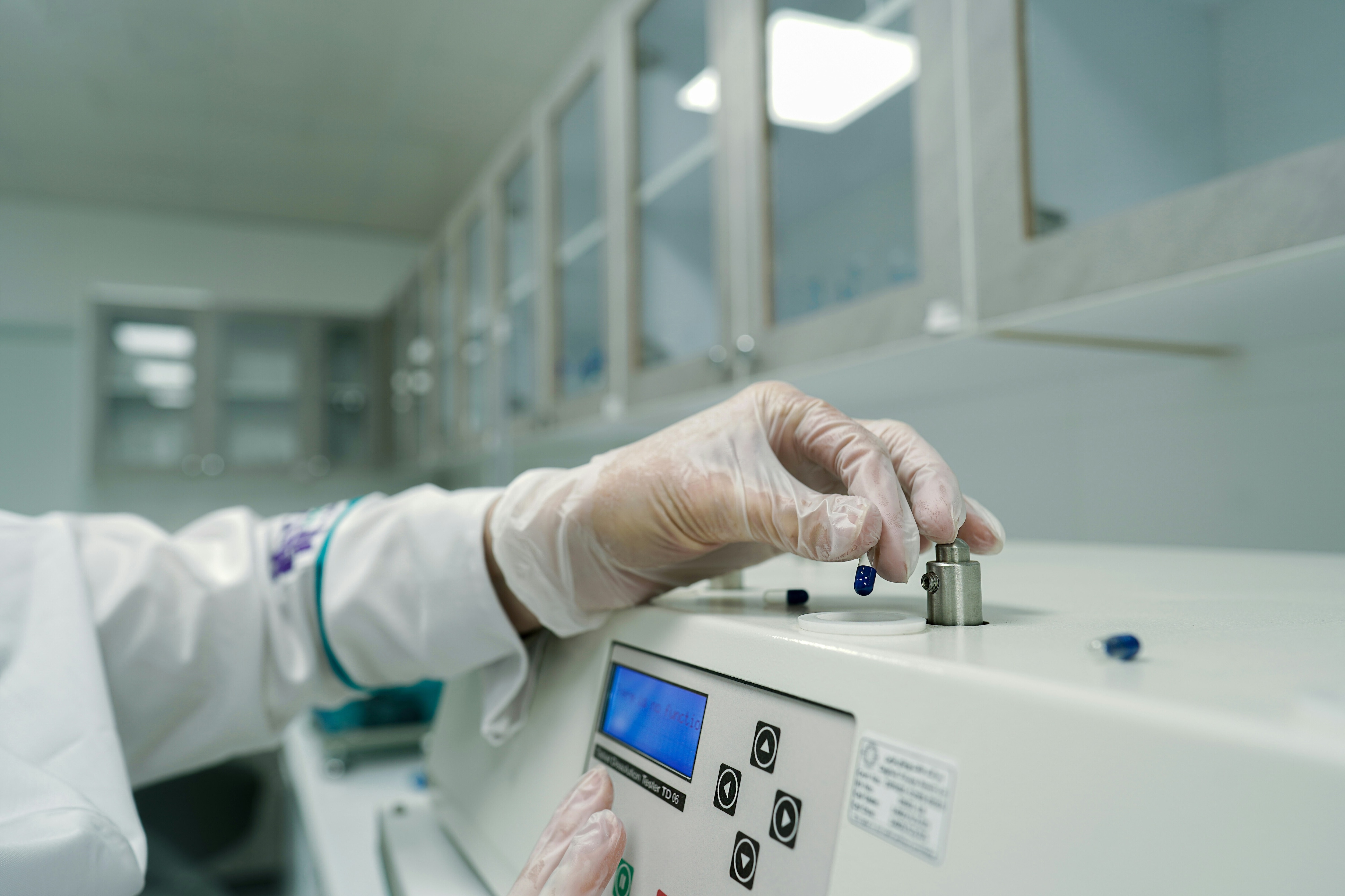 A close-up side view of a technician wearing a white lab coat and blue protective gloves operating a modern, automated laboratory machine with a digital interface.