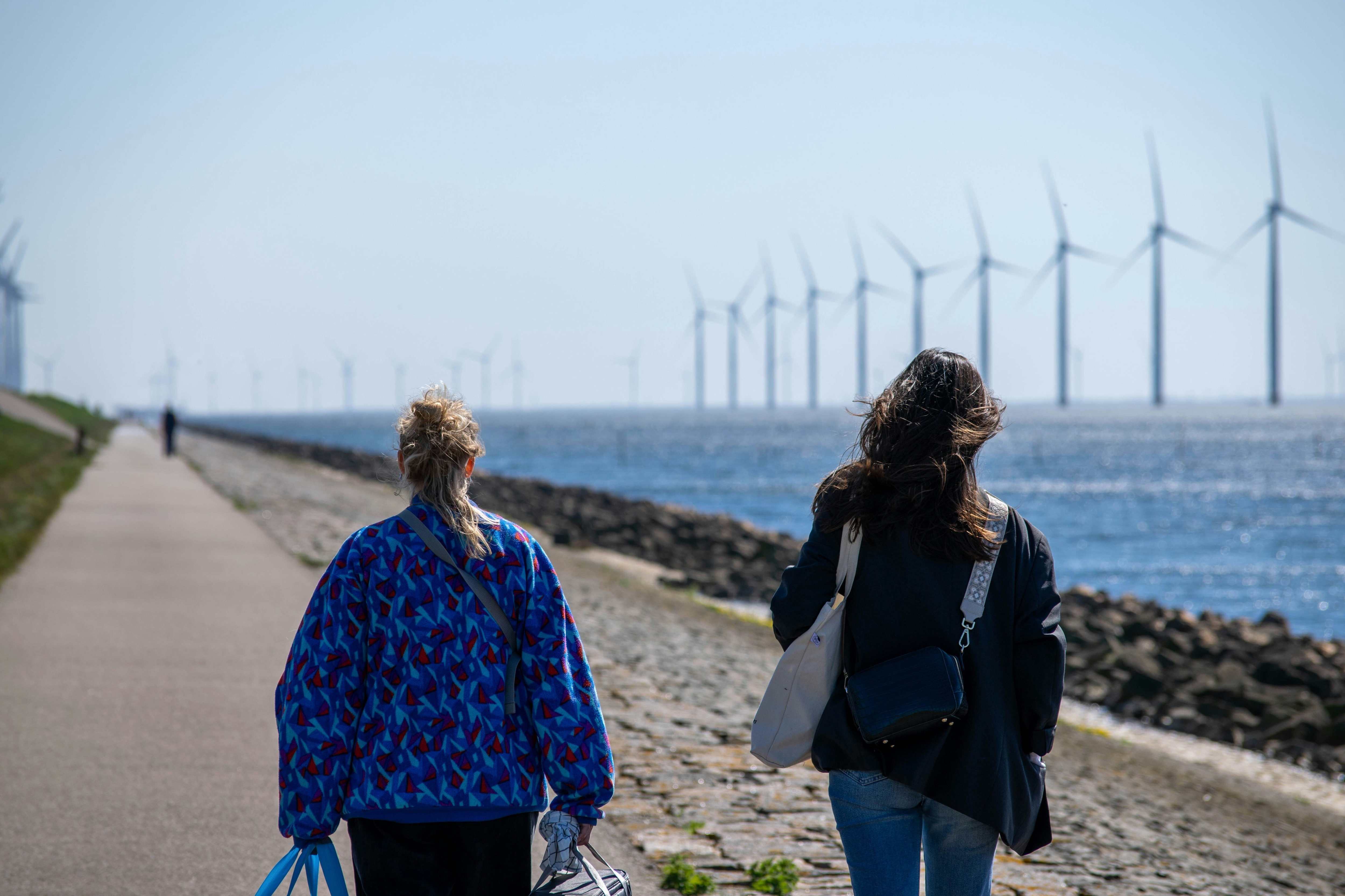 Two women walk along a paved coastline with wind turbines in the distance: AI is both the motivator and key customer for sustainability