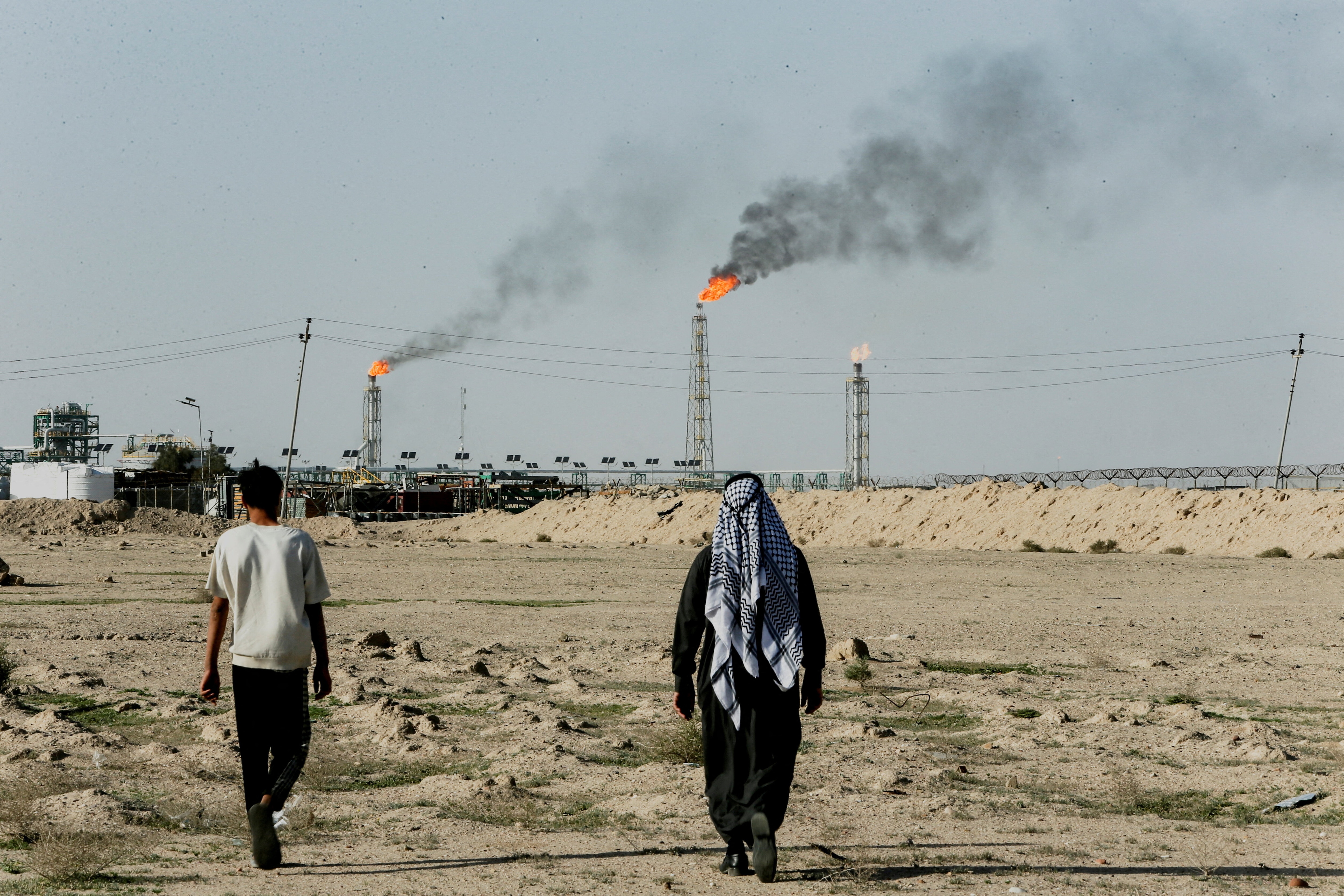 People walk near farmland by the Zubair oil field as gas flares rise in the distance, in Zubair Mishrif, Basra, Iraq.