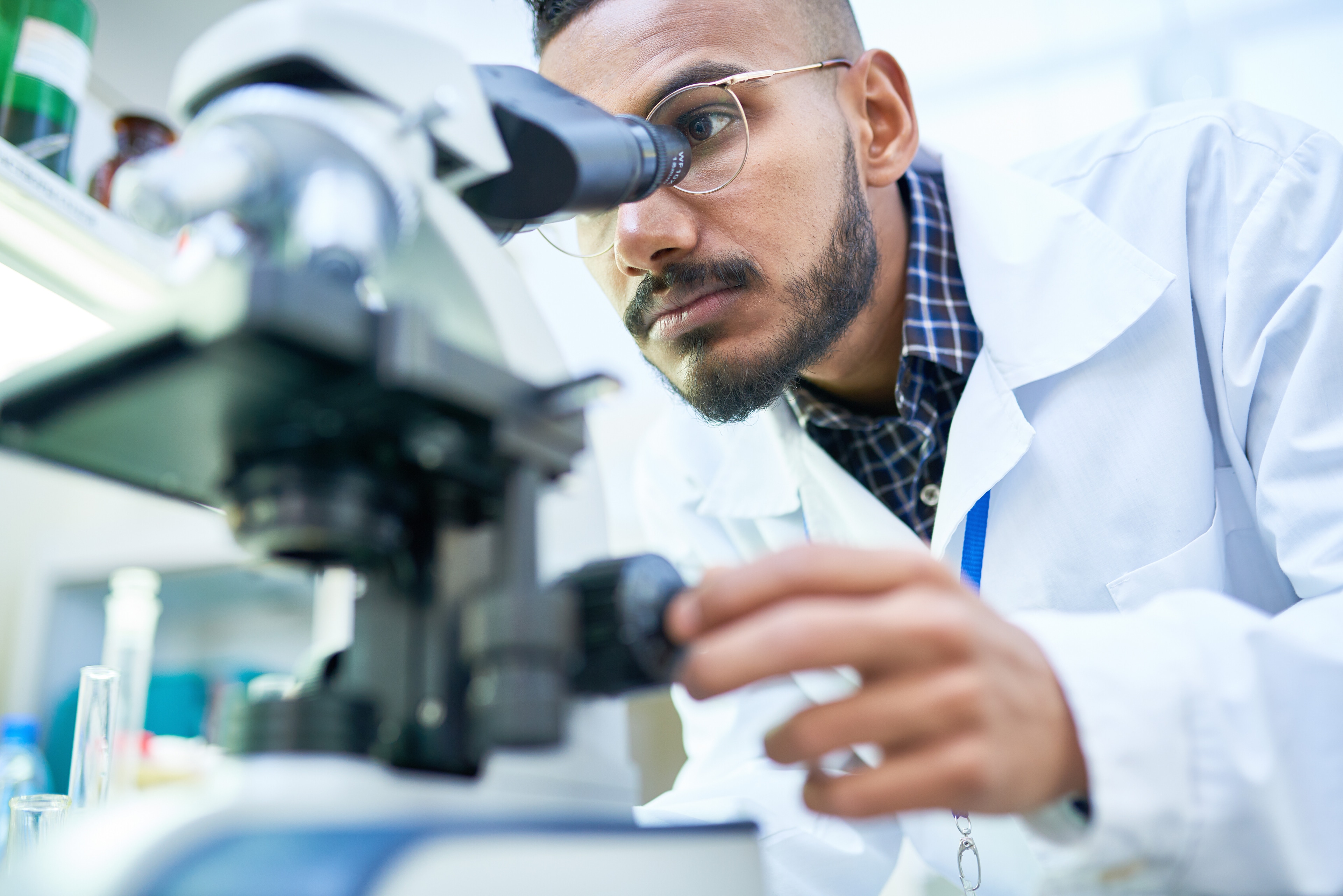 Portrait of young Middle-Eastern scientist looking in microscope while working on medical research in science laboratory, copy space; clinical trial