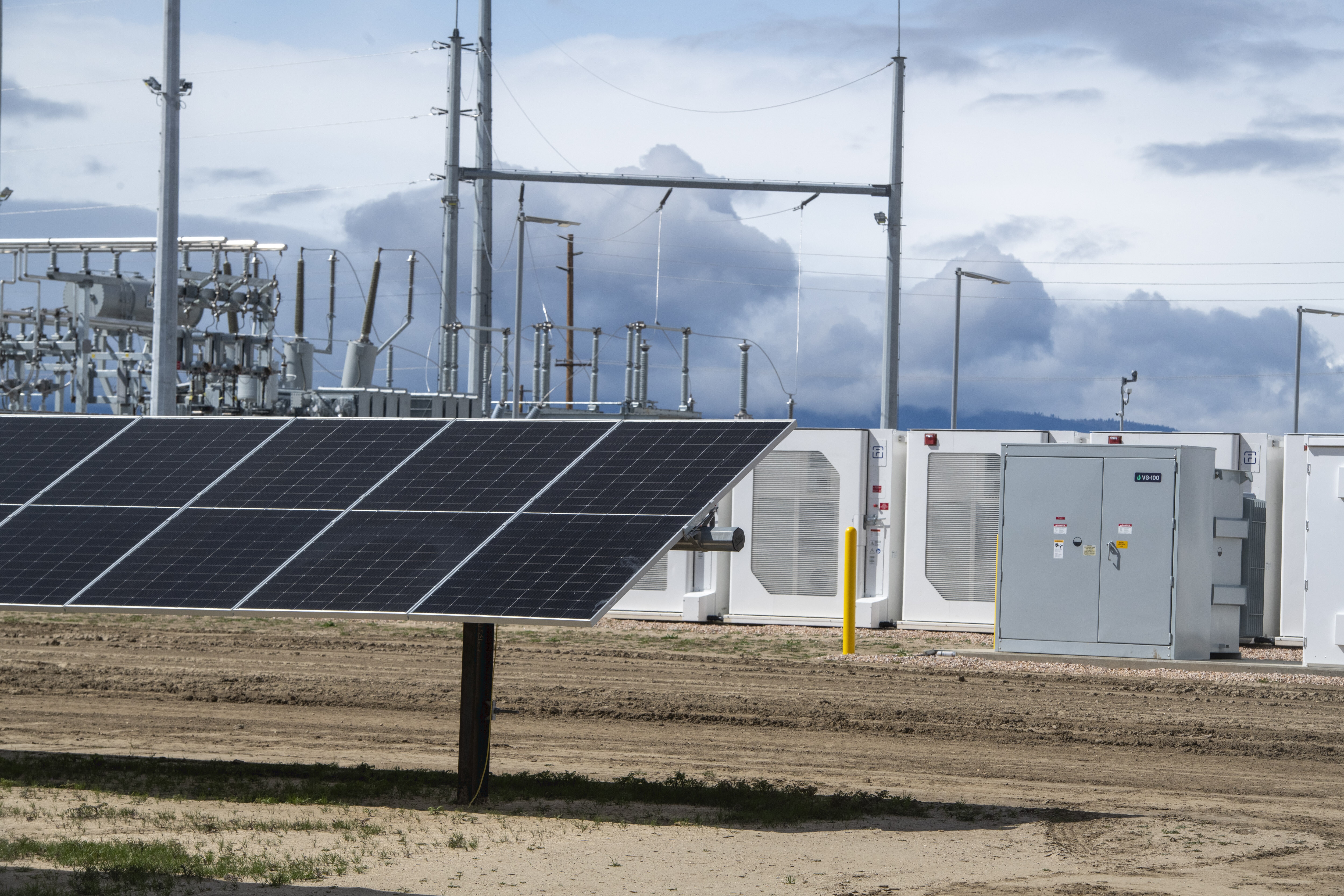 A solar panel in front of infrastructure, including battery storage; sky with clouds in the background