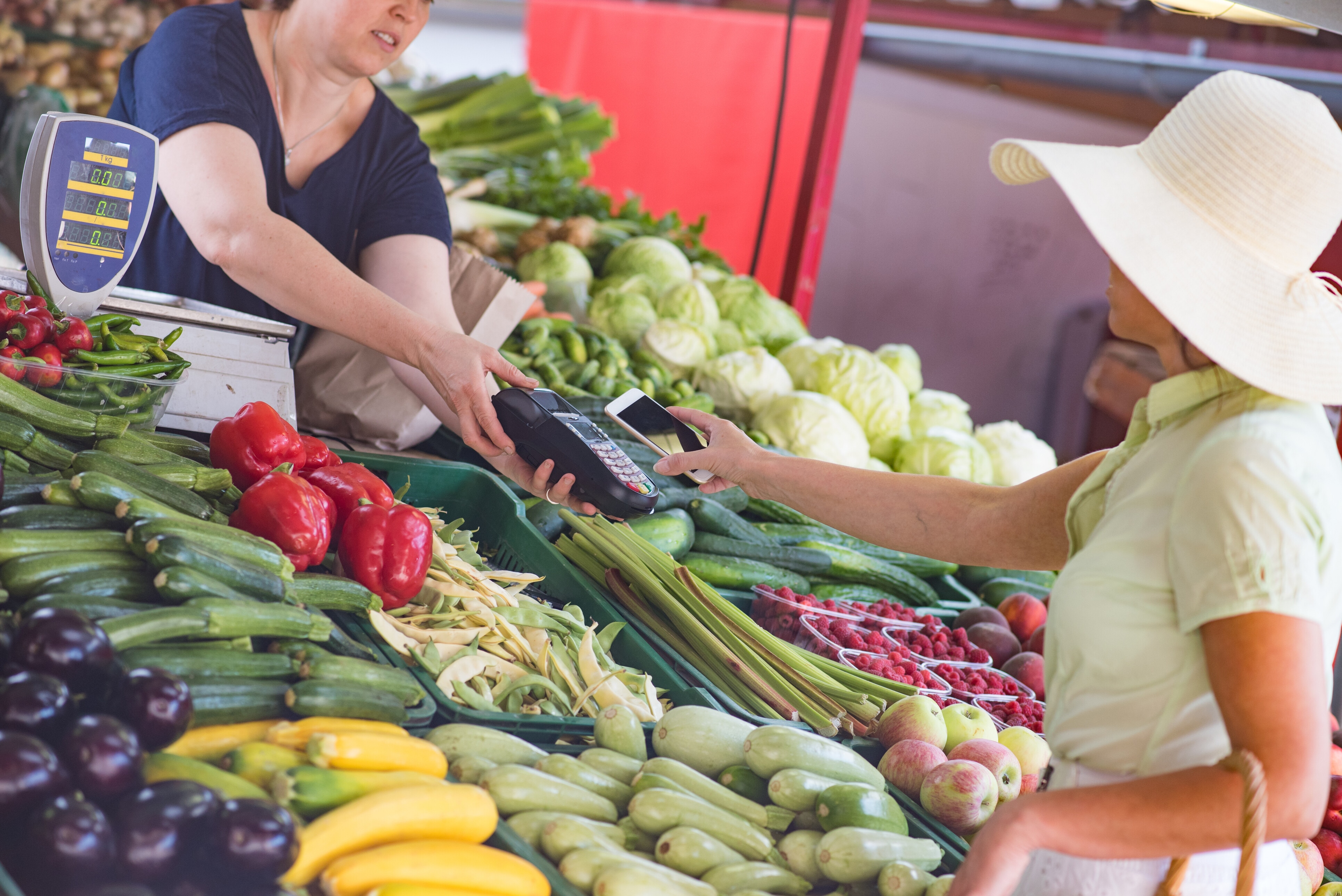 Woman paying for vegetables at market, using credit card reader and smart phone. Digital products