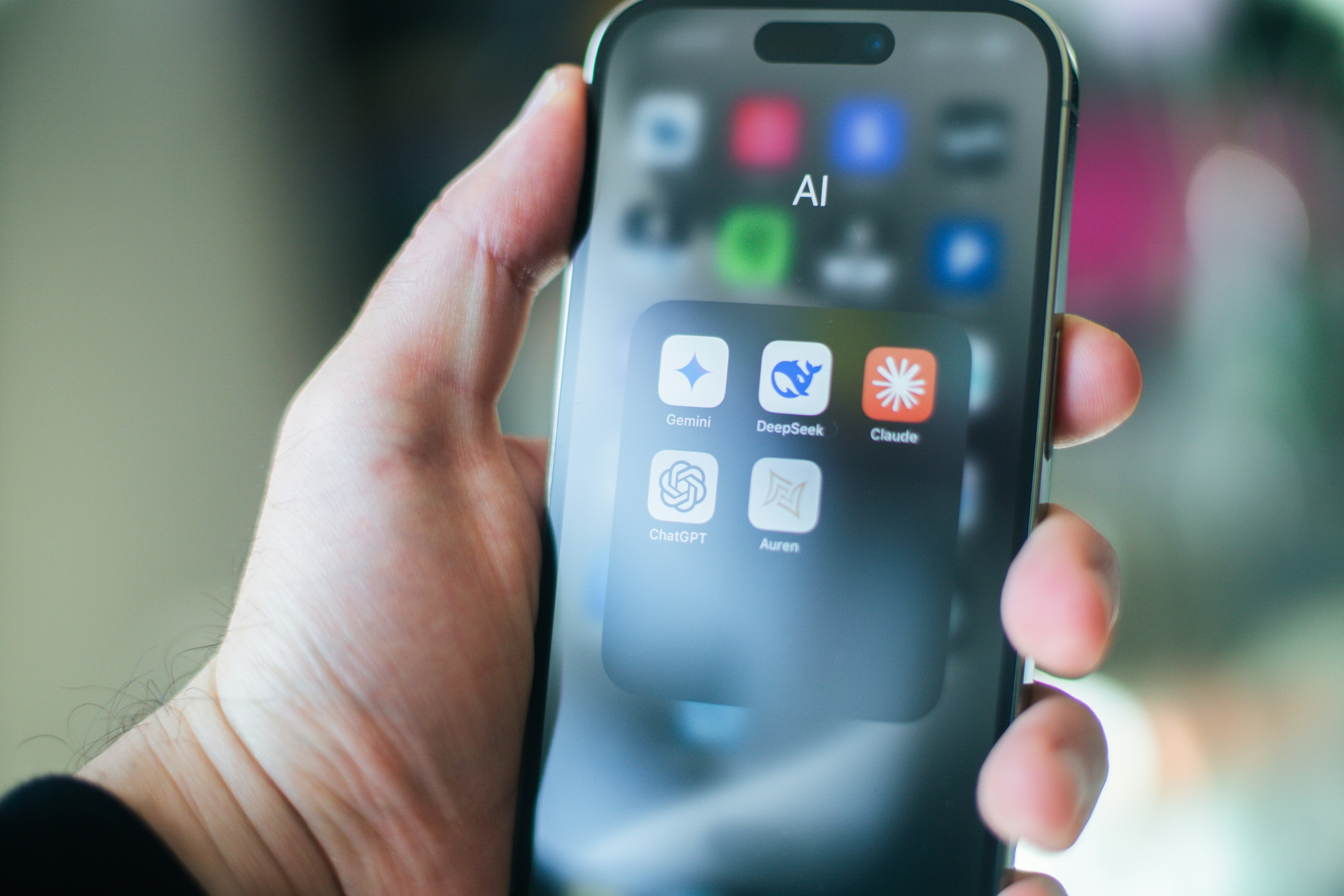 A man's hand holding a smartphone, which shows icons for AI apps including ChatGPT, Claude and Gemini.