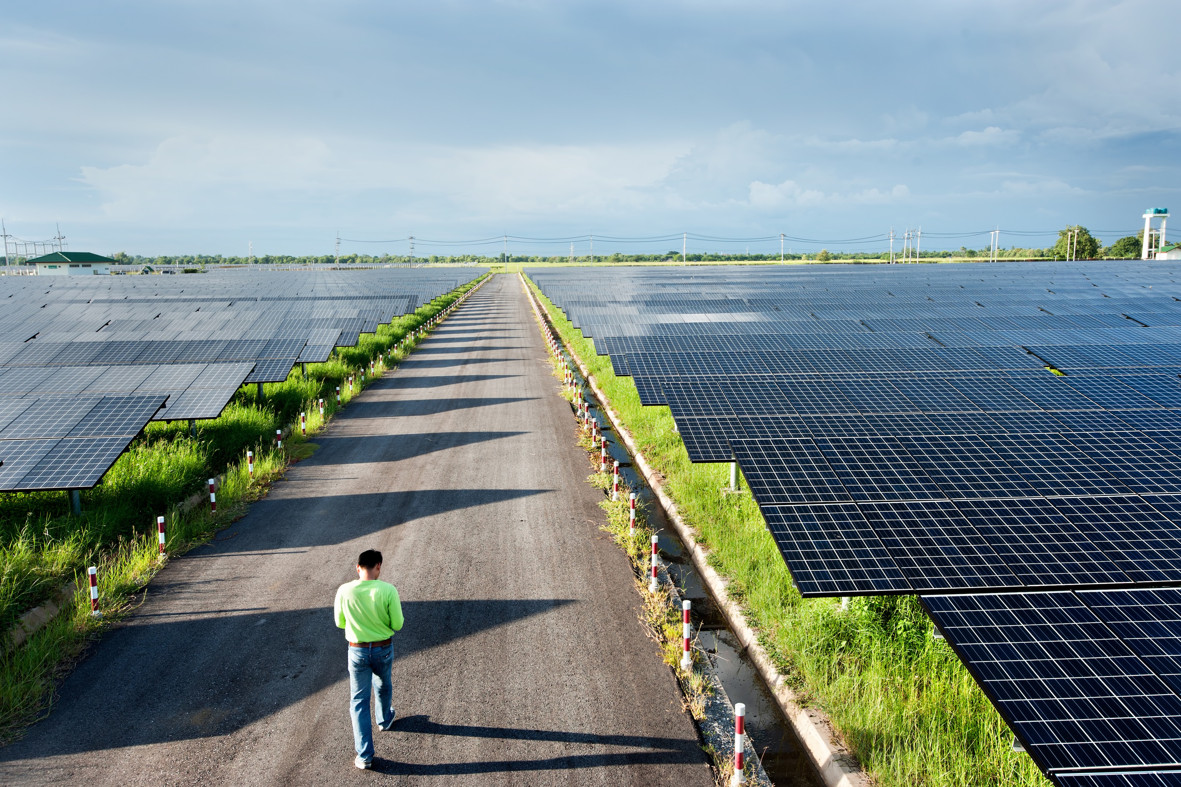 A man walks through a field of solar panels, electricity poles and wires in the background, grass, sky; technology innovation