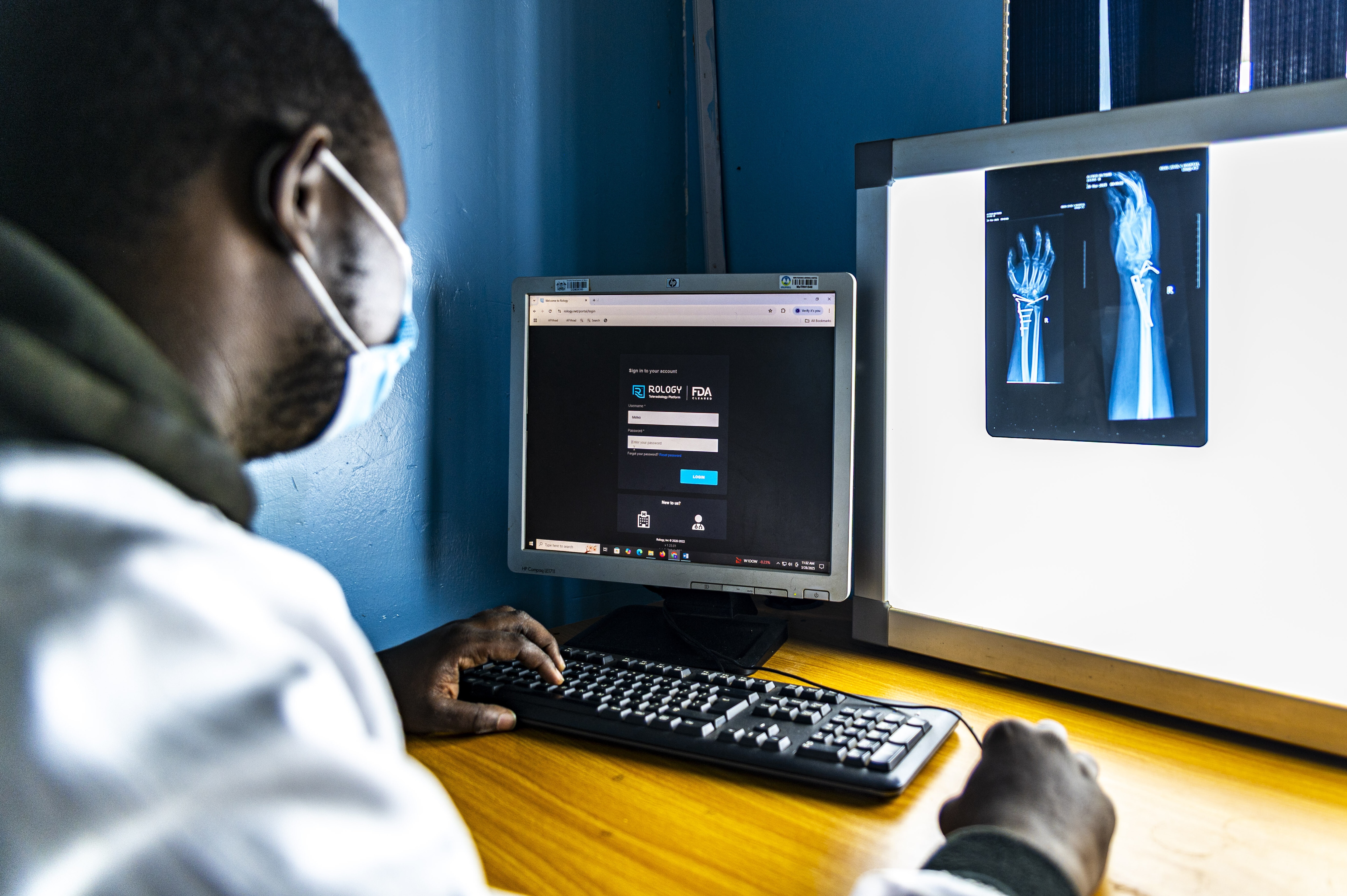 A masked healthcare worker reviews a login screen on a computer and types on a keyboard. An x-ray of an arm is on the wall behind the screen.