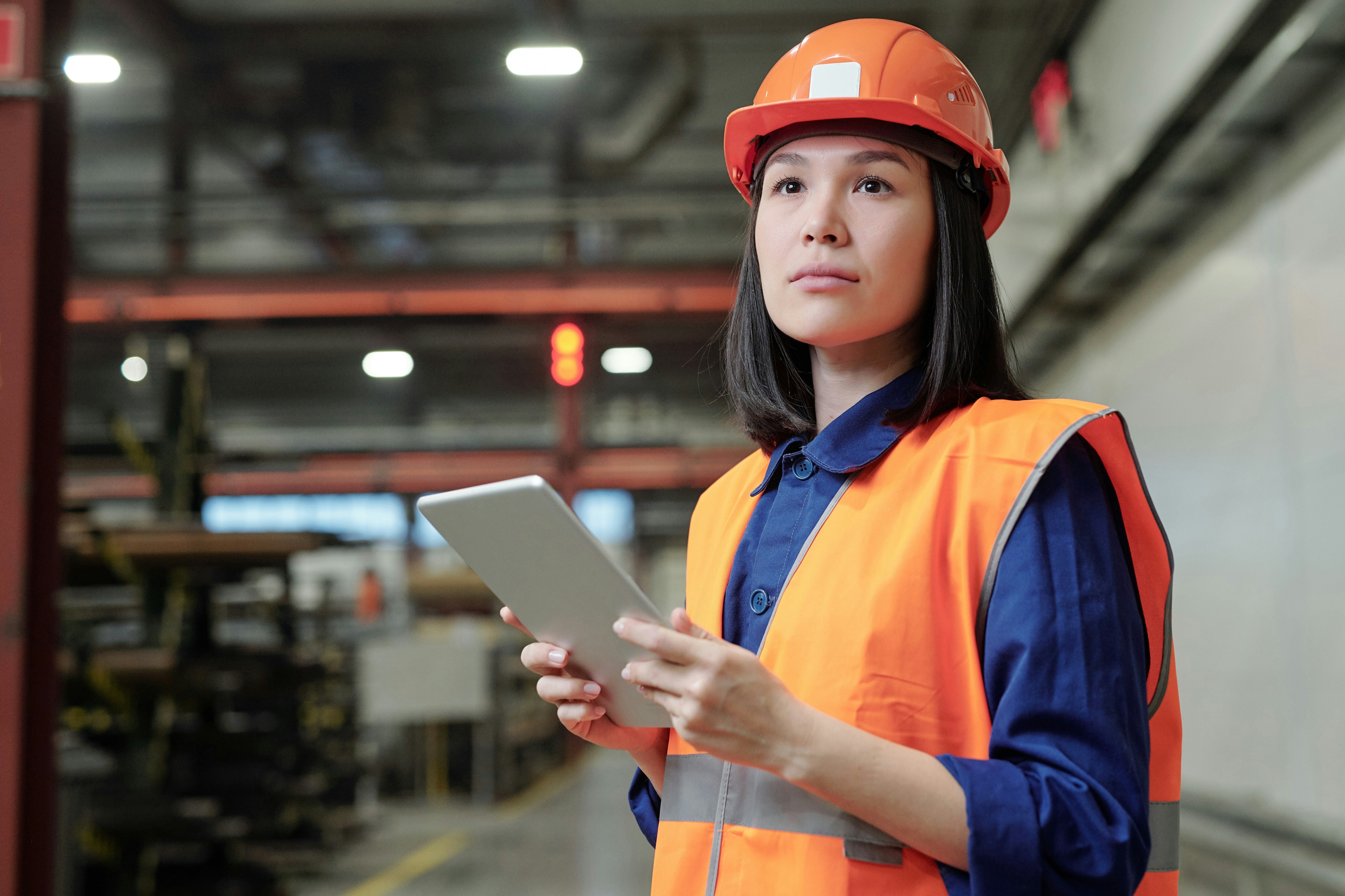 Process engineer in work helmet and safety vest using digital tablet at factory; asian, female; intelligent manufacturing